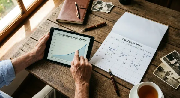 Vanaf boven gefotografeerd: iemands handen volgen een langetermijngrafiek voor pensioenbeleggen op een tablet, die op een bureau ligt naast een kalender die openstaat op een datum ver in de toekomst.