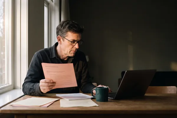 Een man aan een keukentafel leest een afschrift bij het raam, met een laptop en papieren voor zich