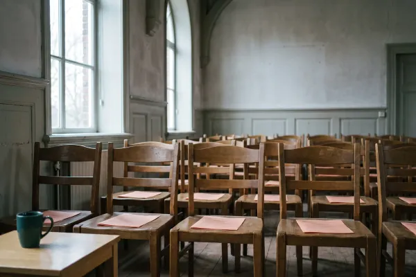 Een lege vergaderzaal met rijen houten stoelen en op elke zitting een vel papier